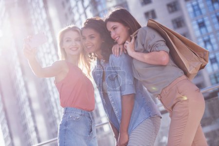 Téléchargez les photos : Belles filles en vêtements décontractés font selfie à l'aide d'un téléphone intelligent, tenant des sacs à provisions et souriant tout en marchant dans la ville - en image libre de droit