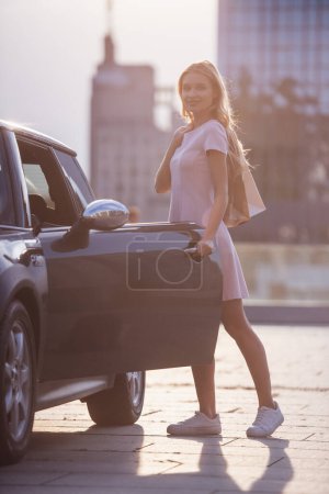 Foto de Hermosa chica rubia en ropa casual se está metiendo en su coche, mirando a la cámara y sonriendo - Imagen libre de derechos