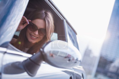 Téléchargez les photos : Belle fille brune regarde dans le miroir latéral et souriant tout en conduisant sa voiture - en image libre de droit