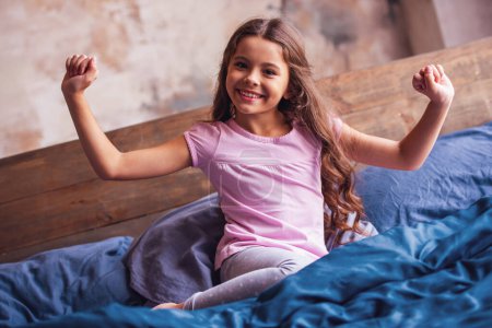 Photo for Pretty little girl is stretching, looking at camera and smiling while lying in bed in the morning - Royalty Free Image