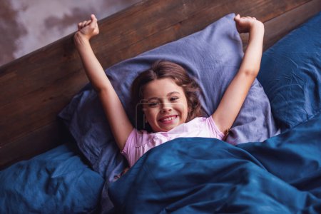 Téléchargez les photos : Jolie petite fille s'étire, regardant la caméra et souriant tout couché dans le lit le matin - en image libre de droit