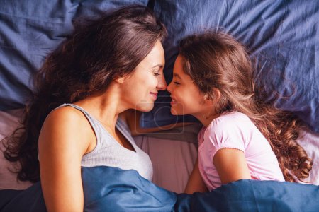 Photo for Beautiful mom and daughter are touching with their noses and smiling while lying in bed at home - Royalty Free Image
