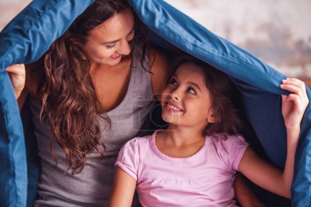Photo for Beautiful mom and daughter are looking at each other and smiling while sitting on bed covered with a duvet - Royalty Free Image