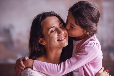 Foto de Hermosa mamá y su hija están abrazando, mamá está sonriendo mientras que la chica la besa en la mejilla - Imagen libre de derechos