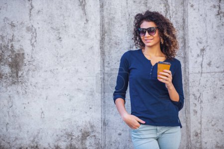 Photo for Attractive girl in casual clothes and sun glasses is holding a cup of coffee, looking at camera and smiling, on concrete wall background - Royalty Free Image