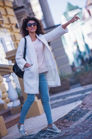 Attractive young woman in casual clothes and sun glasses is holding out her hand and smiling while catching a car in the city