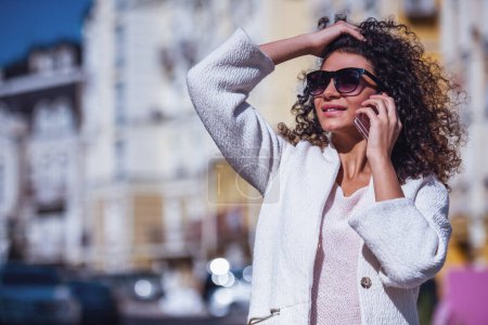 Foto de Mujer joven atractiva en abrigo blanco y gafas de sol está hablando en el teléfono móvil y sonriendo mientras camina por la ciudad - Imagen libre de derechos