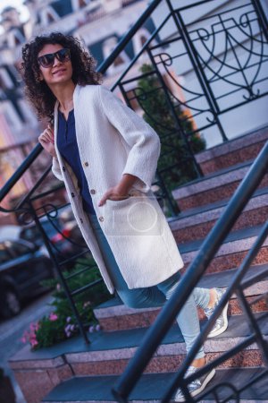 Téléchargez les photos : Jolie jeune femme en manteau blanc et lunettes de soleil sourit tout en marchant dans la ville - en image libre de droit