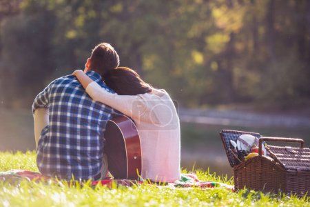 Foto de Vista trasera de la hermosa pareja joven abrazándose mientras descansa en un cuadros en el parque, el chico está tocando la guitarra - Imagen libre de derechos