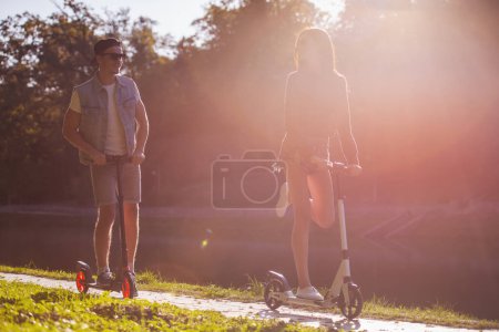 Photo for Stylish young couple is smiling while riding scooter in the park - Royalty Free Image
