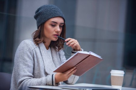 Téléchargez les photos : Belle fille en manteau et chapeau lit le livre tout en travaillant dans un café à l'extérieur - en image libre de droit