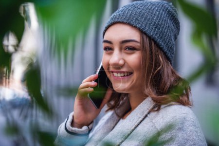 Photo for Beautiful girl in coat and cap is talking on the mobile phone and smiling while resting outdoors - Royalty Free Image