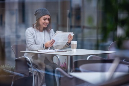 Téléchargez les photos : Belle fille en manteau et casquette lit un journal et sourit tout en se reposant dans un café à l'extérieur - en image libre de droit