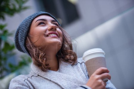 Foto de Hermosa chica con abrigo y gorra está bebiendo café y sonriendo mientras camina al aire libre - Imagen libre de derechos