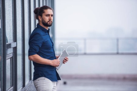 Photo for Stylish young businessman with long dark hair is holding a digital tablet while standing outdoors - Royalty Free Image