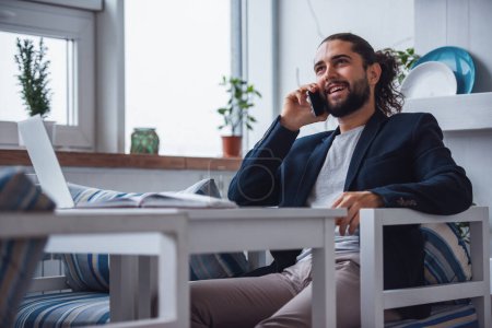 Téléchargez les photos : Beau jeune homme d'affaires aux longs cheveux bruns parle sur le téléphone portable et sourit tout en travaillant dans un café - en image libre de droit