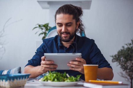 Téléchargez les photos : Beau jeune homme aux longs cheveux noirs utilise une tablette numérique et souriant tout en mangeant dans le café - en image libre de droit