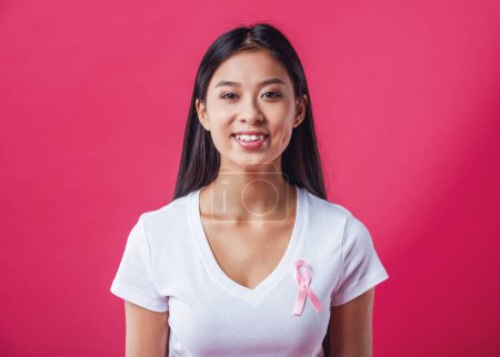 Photo for Women against breast cancer. Beautiful Asian woman with a pink ribbon on her chest is looking at camera and smiling, on red background - Royalty Free Image