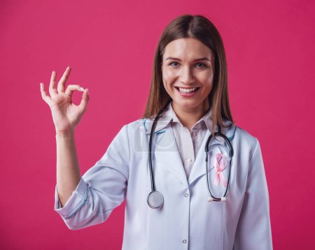 Foto de Mujeres contra el cáncer de mama. Hermosa doctora con una cinta rosa en el pecho está mostrando signo Ok, mirando a la cámara y sonriendo, sobre fondo rojo - Imagen libre de derechos