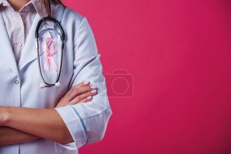 Téléchargez les photos : Femmes contre le cancer du sein. Image recadrée d'une femme médecin avec un ruban rose sur la poitrine debout avec les bras croisés, sur fond rouge - en image libre de droit