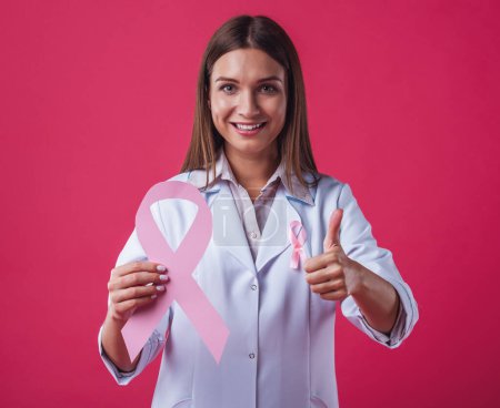 Foto de Mujeres contra el cáncer de mama. Hermosa doctora está sosteniendo una cinta rosa, mostrando signo Ok y sonriendo, sobre fondo rojo - Imagen libre de derechos