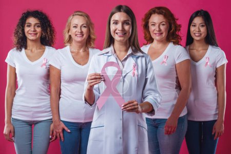 Photo for Women against breast cancer. Beautiful women of different ages and nationalities with pink ribbons on their chests smiling, doctor holding a pink paper ribbon, on red background - Royalty Free Image
