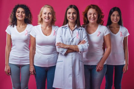 Téléchargez les photos : Femmes contre le cancer du sein. De belles femmes d'âges et de nationalités différents avec des rubans roses sur leur poitrine regardent la caméra et sourient, sur fond rouge - en image libre de droit