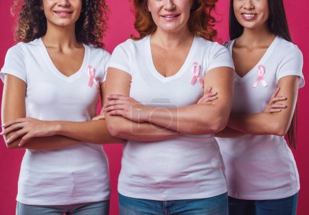 Photo for Women against breast cancer. Beautiful women of different ages and nationalities with pink ribbons on their chests are smiling, cropped, on red background - Royalty Free Image