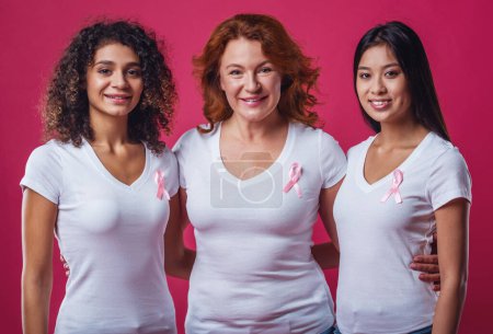 Photo for Women against breast cancer. Beautiful women of different ages and nationalities with pink ribbons on their chests are looking at camera and smiling, on red background - Royalty Free Image