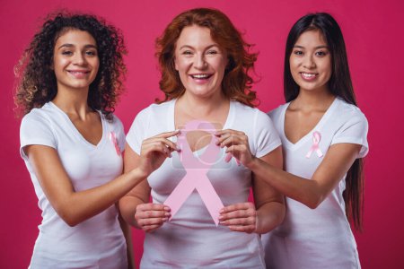 Photo for Women against breast cancer. Beautiful women of different ages and nationalities with pink ribbons on their chests are holding a pink paper ribbon and smiling, on red background - Royalty Free Image