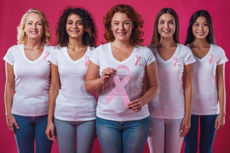 Photo for Women against breast cancer. Beautiful women of different ages and nationalities with pink ribbons on their chests smiling, one is holding a pink paper ribbon, on red background - Royalty Free Image