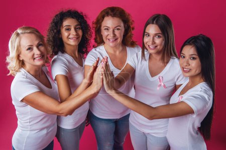 Photo for Women against breast cancer. Beautiful women of different ages and nationalities with pink ribbons on their chests are holding hands together and smiling, on red background - Royalty Free Image
