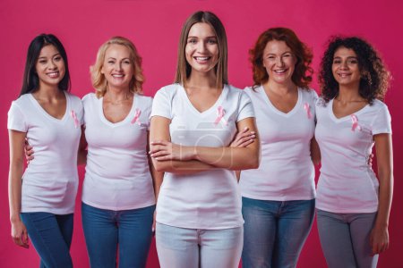 Téléchargez les photos : Femmes contre le cancer du sein. De belles femmes d'âges et de nationalités différents avec des rubans roses sur leurs poitrines sourient, sur fond rouge - en image libre de droit