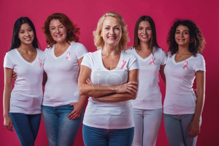 Photo for Women against breast cancer. Beautiful women of different ages and nationalities with pink ribbons on their chests are smiling, on red background - Royalty Free Image