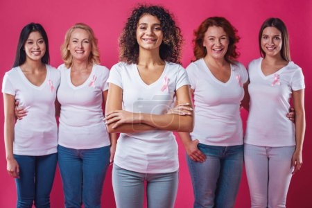 Foto de Mujeres contra el cáncer de mama. Hermosas mujeres de diferentes edades y nacionalidades con cintas rosas en el pecho sonríen, sobre fondo rojo - Imagen libre de derechos