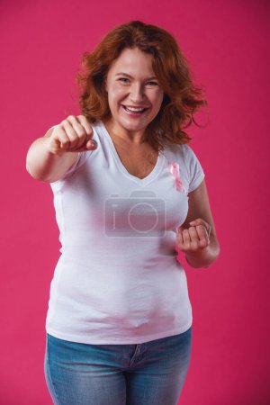 Photo for Women against breast cancer. Beautiful mature woman with pink ribbon on her chest is showing fists and smiling, on red background - Royalty Free Image