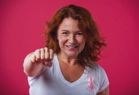 Photo for Women against breast cancer. Beautiful mature woman with pink ribbon on her chest is showing fist and smiling, on red background - Royalty Free Image