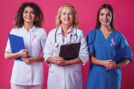 Photo for Women against breast cancer. Beautiful young female doctors with pink ribbons on their chests are holding folders, looking at camera and smiling, on red background - Royalty Free Image