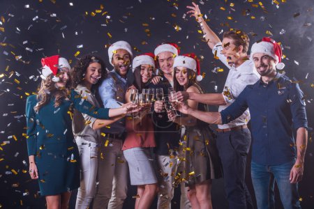 Photo for Happy group of people clinking glasses of champagne together and smiling while celebrating New Year, on dark smoky and with confetti background - Royalty Free Image