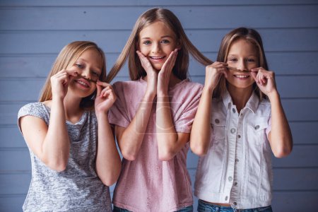 Photo for Three attractive teenage girls in casual clothes are looking at camera and smiling, two are making mustaches of their friend's hair, on gray wall background - Royalty Free Image