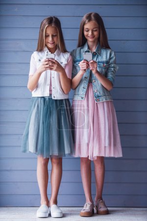 Téléchargez les photos : Portrait pleine longueur de deux adolescentes attrayantes en robes à l'aide de smartphones et souriant, sur fond de mur gris - en image libre de droit