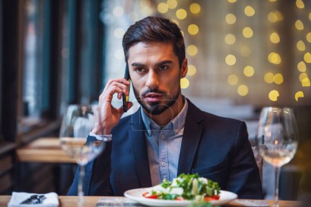 Photo for Handsome businessman in suit is talking on the mobile phone while sitting in restaurant - Royalty Free Image