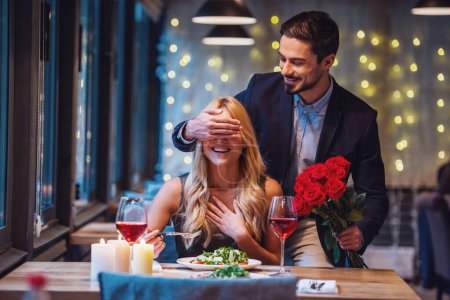 Photo for Handsome elegant man is holding roses and covering his girlfriend's eyes while making a surprise in restaurant, both are smiling - Royalty Free Image