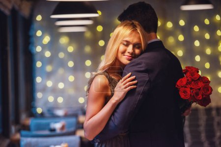 Téléchargez les photos : Belle fille tient des roses et sourit tout en embrassant son petit ami pendant leur rendez-vous au restaurant - en image libre de droit