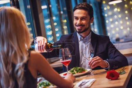 Foto de Hombre elegante guapo está mirando a su novia, vertiendo vino y sonriendo mientras tienen una cita en el restaurante - Imagen libre de derechos