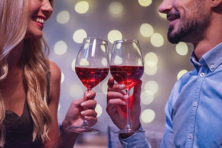 Photo for Beautiful young couple is looking at each other, clinking glasses of wine together and smiling during their date in a restaurant - Royalty Free Image