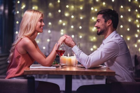 Photo for Beautiful young couple is looking at each other, holding hands and smiling during their date in a restaurant - Royalty Free Image