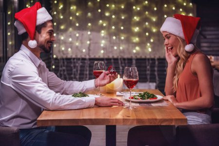 Foto de Hombre guapo sostiene un anillo de compromiso y le propone matrimonio a su novia mientras están vestidos con sombreros de Papá Noel celebrando el Año Nuevo en un restaurante - Imagen libre de derechos