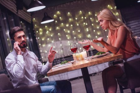 Foto de Hermosa joven mujer está molesta mientras su novio está hablando en el teléfono móvil durante su cita en un restaurante - Imagen libre de derechos