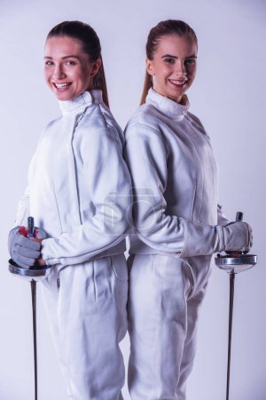 Photo for Beautiful female fencers in protective clothing are holding weapon, looking at camera and smiling, on gray background - Royalty Free Image